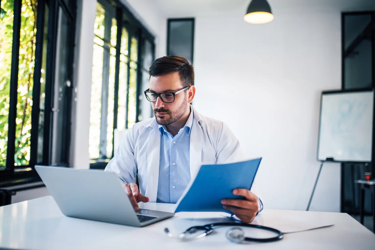A man in a white coat sits at a desk, working on a laptop.