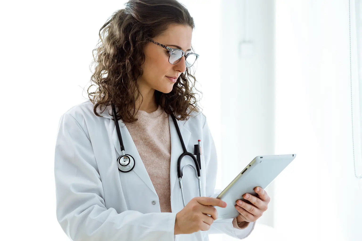 A female doctor in a white coat is focused on using a tablet in a clinical setting.