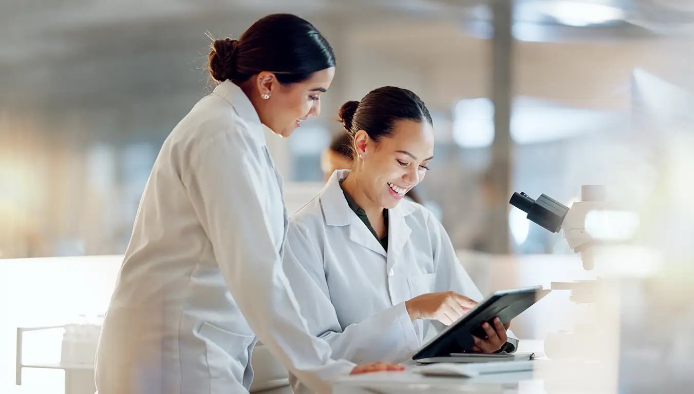 Two women in lab coats examine a tablet together, discussing data in a laboratory setting.